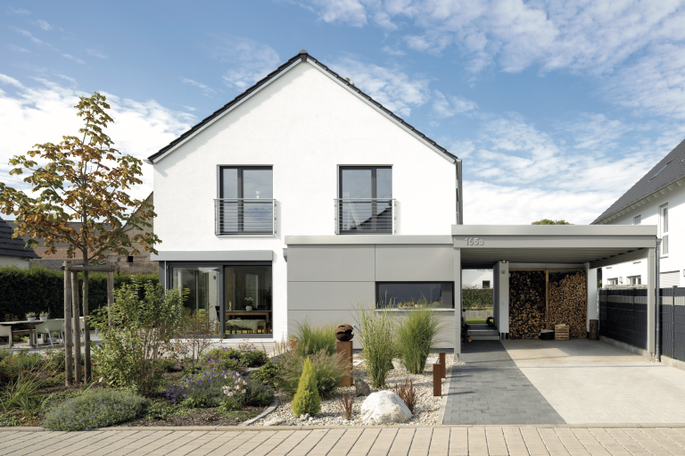 Regnauer zeigt ein weisses Einfamilienhaus mit grauen Akzenten und mit einem Wintergarten, einer gefliesten Terrasse mit einer Sitzgruppe aus Holz und einem grauen Carport in dem Brennholz gelagert wird.