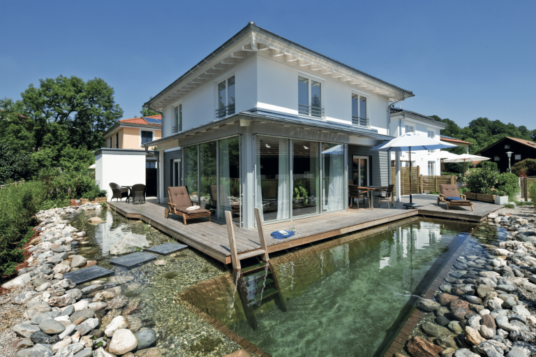 Regnauer zeigt ein modernes Einfamilienhaus mit Flachdach und großen Glasfronten und einem Garten mit Schwimmteich und einem Sonnendeck aus Holz und gemütlicher Liege mit brauner Polsterauflage.