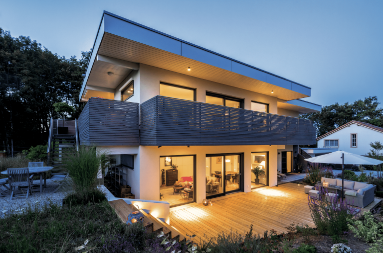 Regnauer zeigt ein modernes Einfamilienhaus mit Flachdach, großen Glasfronten und einem Balkon der sich über das ganze Haus erstreckt und einer Terrasse mit Holzdielen und gemütlicher Outdoorcouch mit Sonnenschirm.