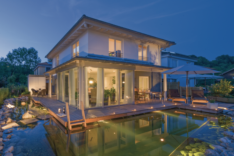 Regnauer zeigt ein modernes Einfamilienhaus mit Flachdach und großen Glasfronten und einem Garten mit Schwimmteich und einem Sonnendeck aus Holz, gemütlichen Liegen und schöner Beleuchtung.