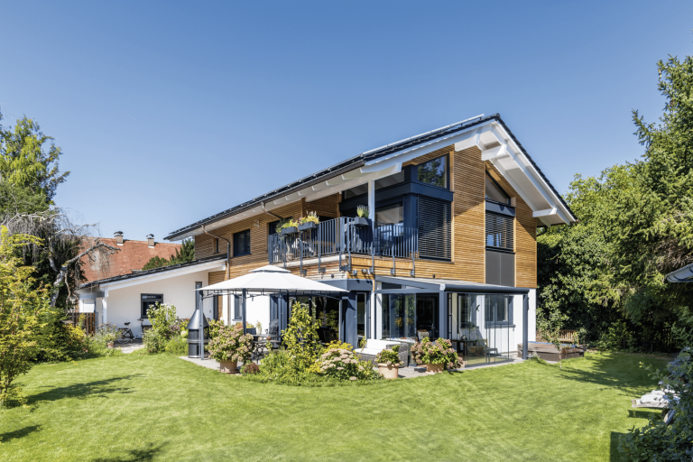 Regnauer zeigt ein Einfamilienhaus mit Satteldach, Holzverbau im ersten Stock, Terrasse, Terrassenüberdachung, Sonnenschutz und Garten.