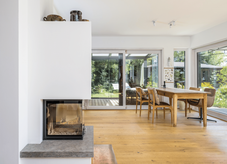 Regnauer zeigt ein Esszimmer mit Holzboden, Esstisch und Stühle ebenfalls aus Holz, einen eingemauerten Ofen und Schiebefenster, welche auf die Terrasse führen.