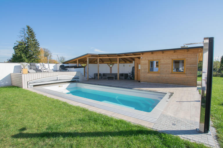 Friedrich Reitinger Schwimmbad und Wellness zeigt einen in den Boden eingelassenen, rechteckigen Pool in einem gepflegten Garten mit einer Poolüberdachung und einem Sonnendeck.