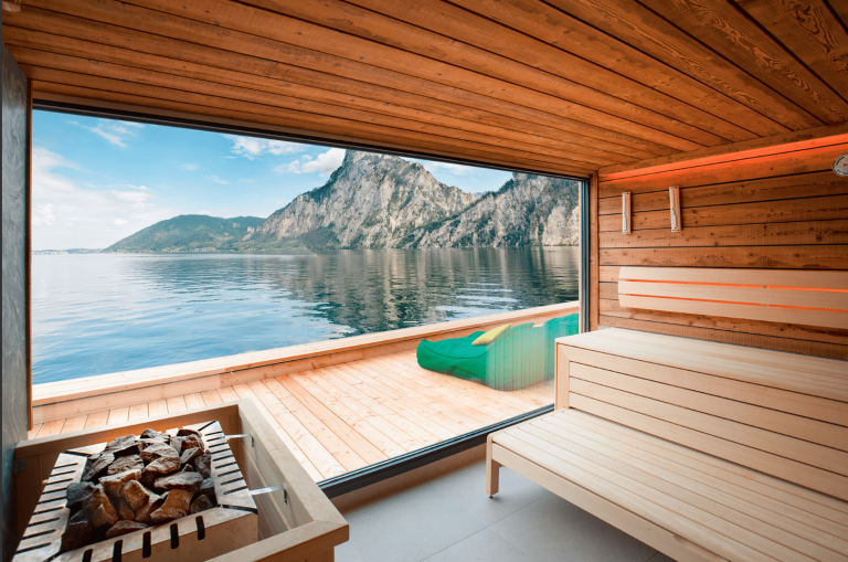 SAUNA-SCHWIMMBAD RUHA Stelzmüller zeigt eine Sauna mit Holzverkleidung und beeindruckendem Seeblick durch ein Panoramafenster.