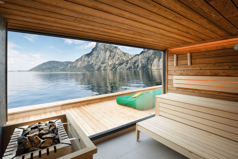 RUHA präsentiert eine Sauna aus Holz von innen, mit dem Aufgussblock vor einem Fenster mit Blick in die Berge und einer Terrasse mit Liegen.