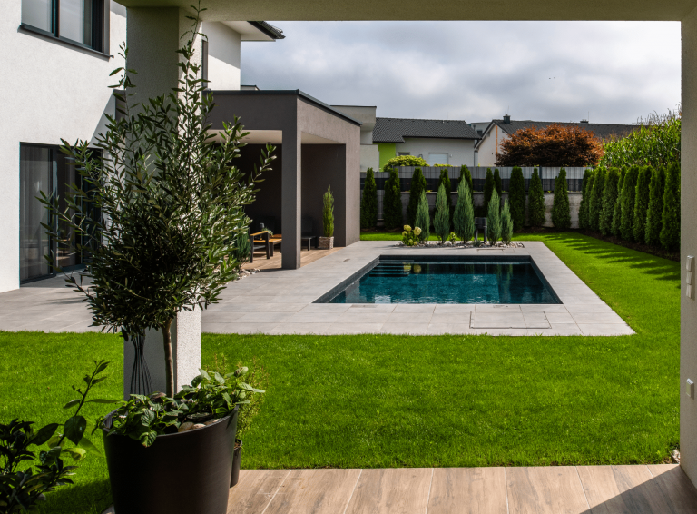 RUHA zeigt einen edlen dunklen Pool der im Boden versenkt ist und eine geflieste Umrandung hat im gepflegten Garten von einem Einfamilienhaus mit überdachter Terrasse.