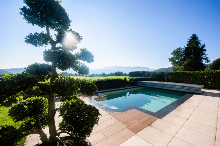 RUHA zeigt einen rechteckigen Pool mit rundem Bereich für den Einstieg, Stufen und einer Poolüberdachung aus Glas zum Schieben auf einer gefliesten Terrasse.