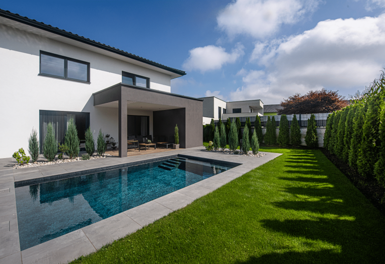 RUHA zeigt einen dunkel gefliesten Pool der im Boden versenkt ist und eine graue Umrandung hat im gepflegten Garten von einem Einfamilienhaus mit überdachter Terrasse.