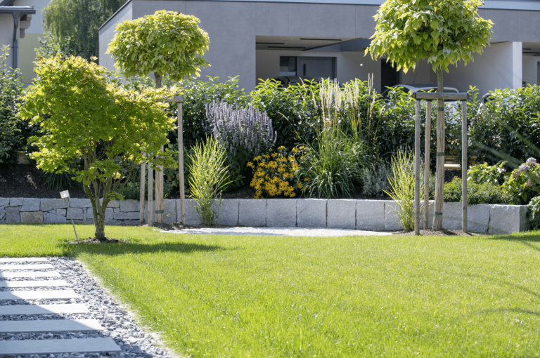 Gartenbau Schmid zeigt ein bepflanztes Beet aus Steinen mit dichter Hecke dahinter als Sichtschutz, angelegten Weg und gepflegtem Rasen.