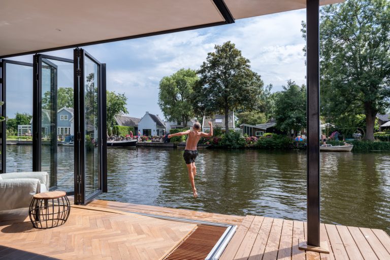 Solarlux zeigt ein Wohnzimmer mit Glasfaltwand zum Holzsteg wodurch man direkt auf ein Boot oder in den Fluss gelangt.