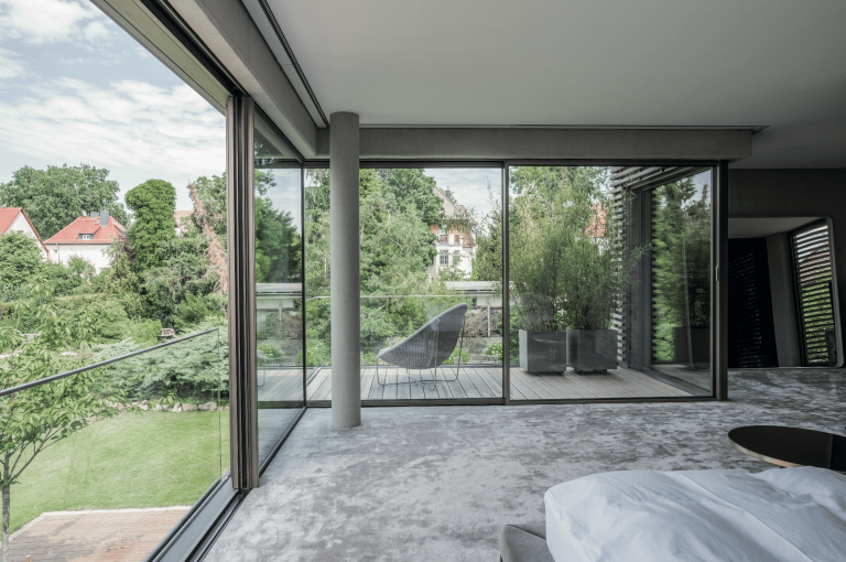 Solarlux zeigt Schlafzimmer mit vollverglasten Fronten mit bodentiefen Fenstern und Glasschiebetür auf den Balkon.