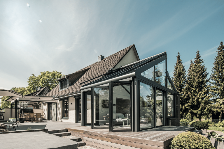 Solarlux zeigt ein Einfamilienhaus mit einer überdachten Terrasse und einen gemütlichen Wintergarten mit Zugang durch eine Glasfaltwand.