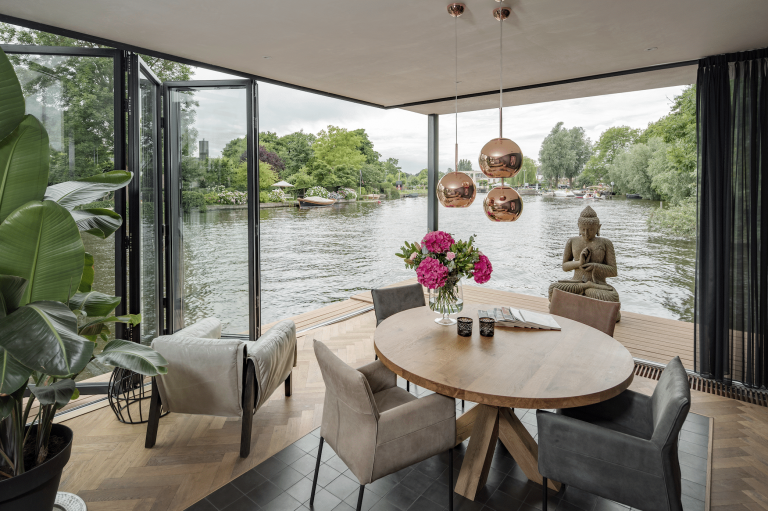 Solarlux zeigt ein Hausboot mit einem gemütlichen Loungebereich, hellem Holzboden und einer Glasfaltwand um die Aussicht auf das Wasser zu genießen.