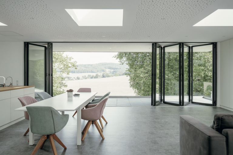 Solarlux zeigt einen offenen Wohnbereich mit Küche, Essbereich mit einem weissen Tisch und Stühlen in Pastelltönen mit einer Glasfaltwand und überdachtem Balkon mit Aussicht ins Grüne.