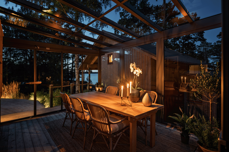 Solarlux zeigt einen schönen Wintergarten mit Glasdach und Glasschiebetüre mit einer Sitzgruppe aus Holz mit Rattanstühlen und gemütlicher Beleuchtung.