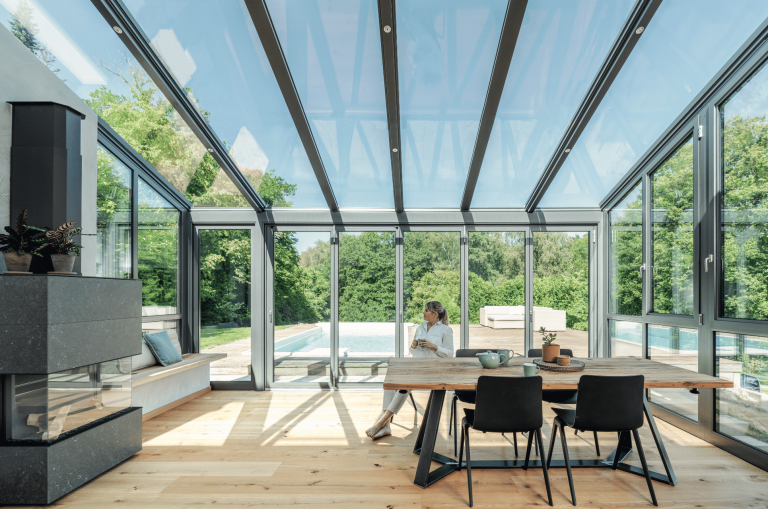 Solarlux zeigt einen großen Wintergarten mit hellem Holzboden, Kamin mit einer Sitzbank daneben und Glasdach, großen Fenstern und einer Glasschiebetür mit Zugang zu dem Garten mit Pool.