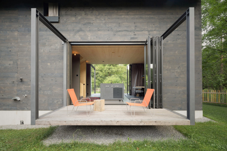 Solarlux zeigt modernes Holzhaus mit Terrasse, seitlichen Glaswänden und Faltschiebetüren mit dunkelgrauem Rahmen mit Blick auf den Garten.