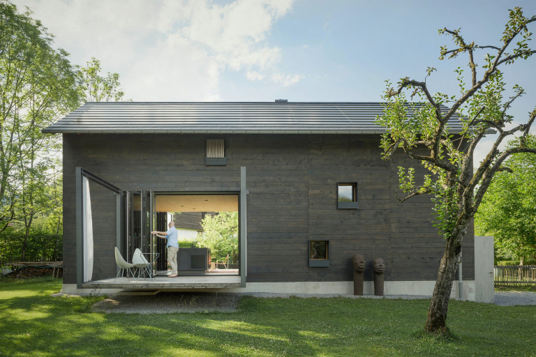 Solarlux zeigt Holzhaus mit Terrasse und Faltschiebetüren mit dunkelgrauem Rahmen, die ein Mann gerade öffnet, mit Blick auf den Garten.