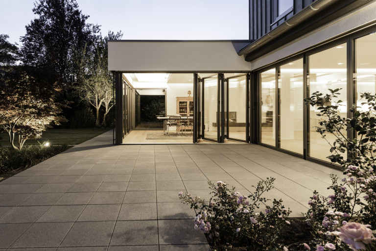 Solarlux zeigt offenes Esszimmer mit halb geöffneten Faltschiebetüren mit dunkelgrauem Rahmen mit Blick auf den Garten bei Abendstimmung.