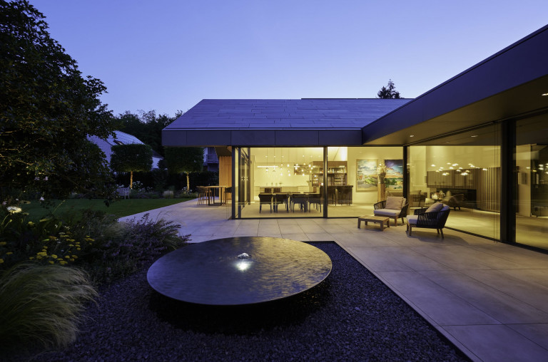 Moderner Bungalow mit Satteldach und bodenbündigen Glasschiebetüren von Solarlux mit dunkelgrauem Rahmen mit Blick auf Terrasse und Garten bei Abendstimmung.