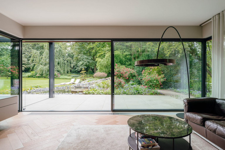 Solarlux zeigt bodenbündige Glasschiebetür mit dunkelgrauem Rahmen mit Blick auf Terrasse und Naturteich.