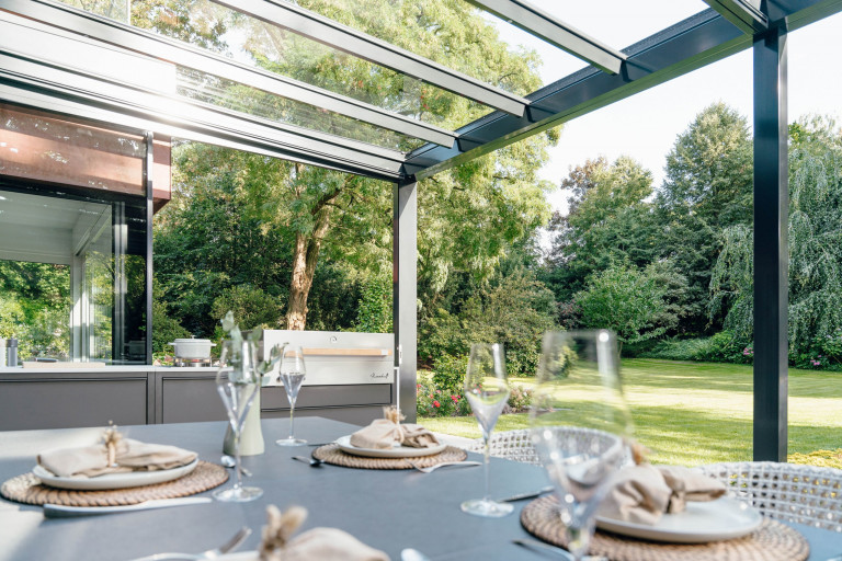 Solarlux zeigt Outdoorküche und Esstisch im Wintergarten aus Alu und Glas mit Glasschiebetüren mit Blick in den Garten.