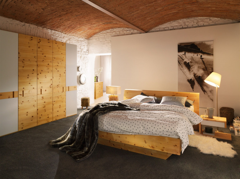 BAUER Tischlerei zeigt gemütliches Schlafzimmer mit Bett aus Kiefer-Holz, passendem Nachttisch und einer Schrankwand mit Kieferholz und weißen Fronten von ANREI in einem Raum mit Gewölbe.