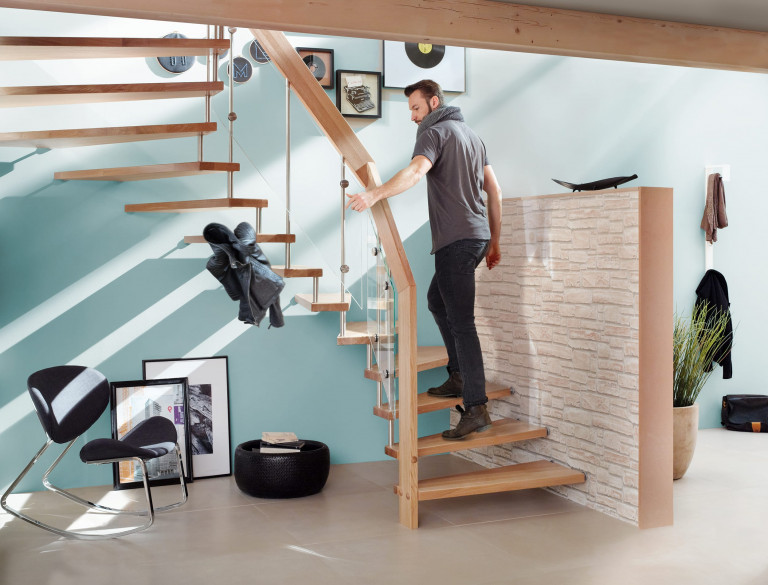 Treppenmeister zeigt freiliegende gedrehte Holztreppe mit Holz-Handlauf und Geländer aus Metallstäben und Glasplatten an einer mintfarbenen Wand.