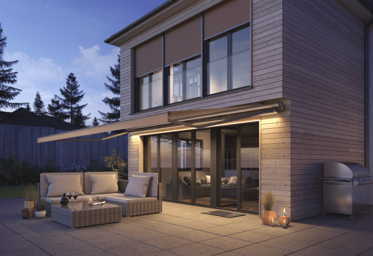 Warema zeigt ein modernes Haus mit beleuchteter Markise über gefliester Terrasse und heller Rattangarnitur für den Outdoorbereich.