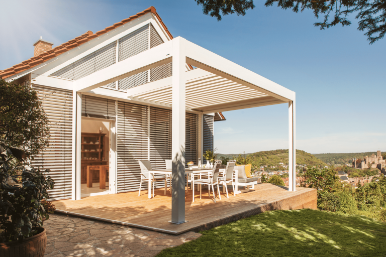 WAREMA zeigt ein weisses Einfamilienhaus mit rotem Spitzdach und einer großen Terrasse mit Holzboden die von einer weissen Pergola überdacht wird und Rollläden an den Fenstern als Sonnenschutz.