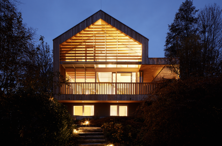 Weissenseer Holz-System-Bau zeigt ein modernes Einfamilienhaus aus Holz mit schöner Beleuchtung, einem Balkon mit Holzgeländer und überdachtem Bereich und vielen Fenstern.