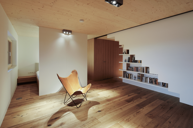 Weissenseer Holz-System-Bau zeigt ein minimalistisches Lesezimmer mit einem Lederstuhl, schönem Holzboden und einem Bücherregal unter der Treppe.