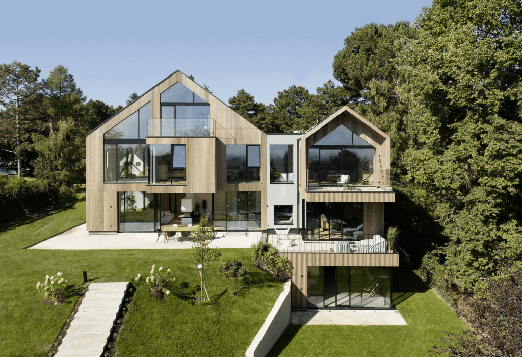 Weissenseer Holz-System-Bau zeigt modernes Wohnhaus mit Holzfassade, doppeltem Satteldach, Mittelbau mit Flachdach, grossen Glasfenstern, Balkon und Terrasse mit Blick auf den Garten.