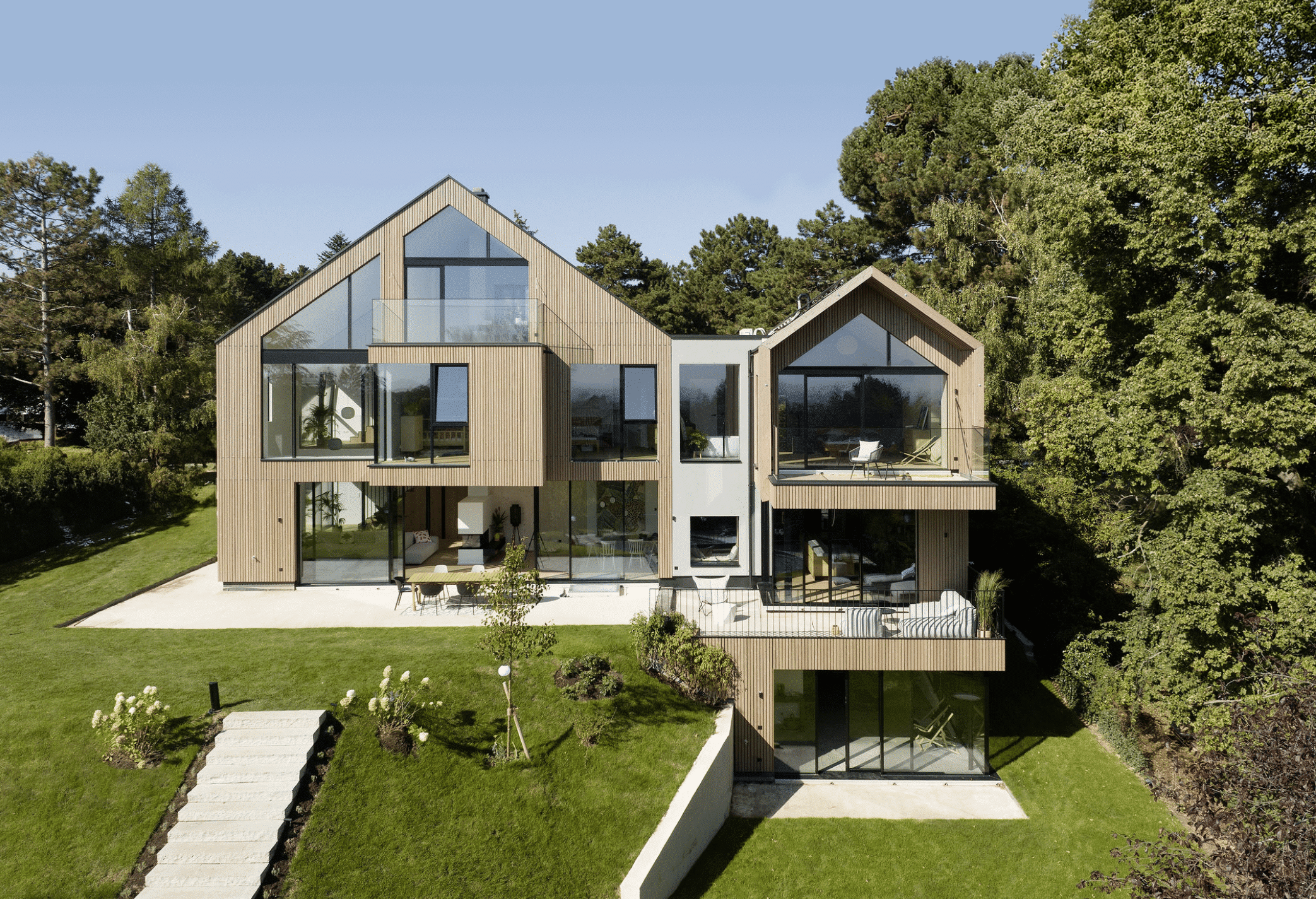 Weissenseer Holz-System-Bau zeigt modernes Wohnhaus mit Holzfassade, doppeltem Satteldach, Mittelbau mit Flachdach, grossen Glasfenstern, Balkon und Terrasse mit Blick auf den Garten.