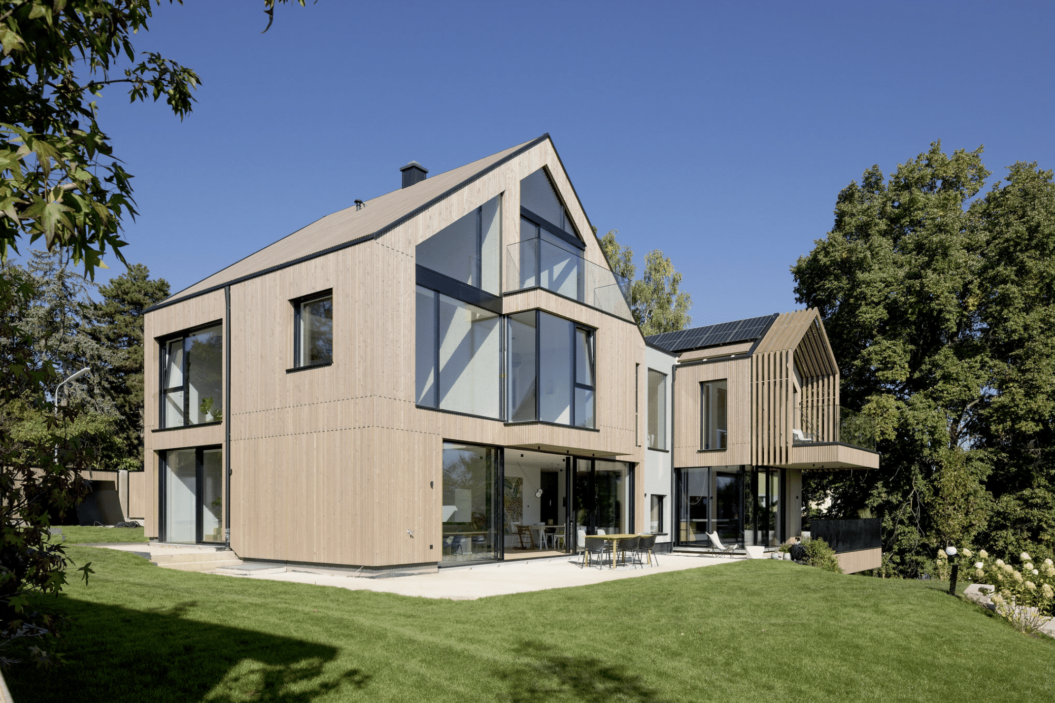 Weissenseer Holz-System-Bau zeigt modernes Wohnhaus mit Holzfassade, doppeltem Satteldach, Mittelbau mit Flachdach, grossen Glasfenstern, Balkon und Terrasse mit Blick auf den Garten.