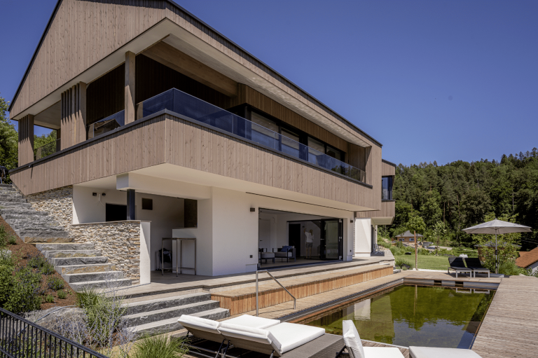 Villa mit Satteldach, Holzfassade, seitlichem Aussenaufgang, Rundum-Balkon, überdachter Terrasse und Naturpool von Weissenseer Holz-System-Bau.