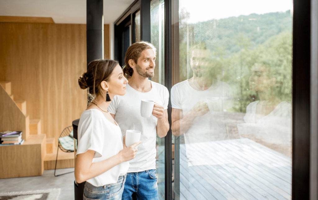 Junges Paar steht mit Kaffetassen im innenraum des neuen Hauses und blickt freudig aus der großflächigen Fensterverglasung in den Garten.