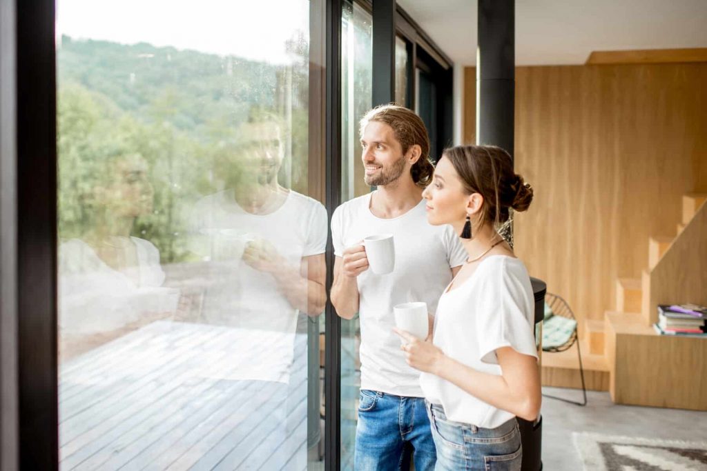 Junges Paar steht mit Kaffetassen im innenraum des neuen Hauses und blickt freudig aus der großflächigen Fensterverglasung in den Garten.